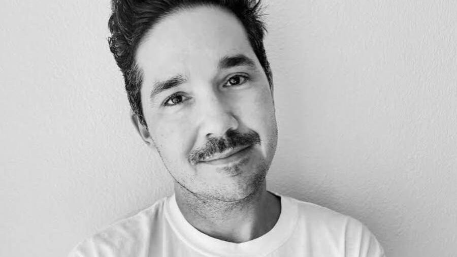 Amos Mac sonriente con una camisa blanca con un rostro gráfico y tatuajes visibles en los antebrazos.