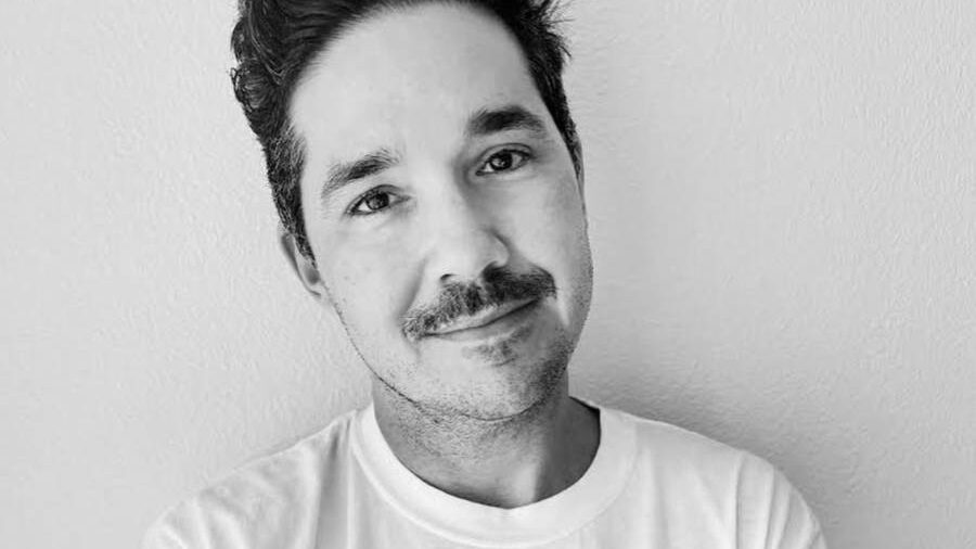 Smiling Amos Mac in a white shirt featuring a graphic face, visible tattoos on forearms.