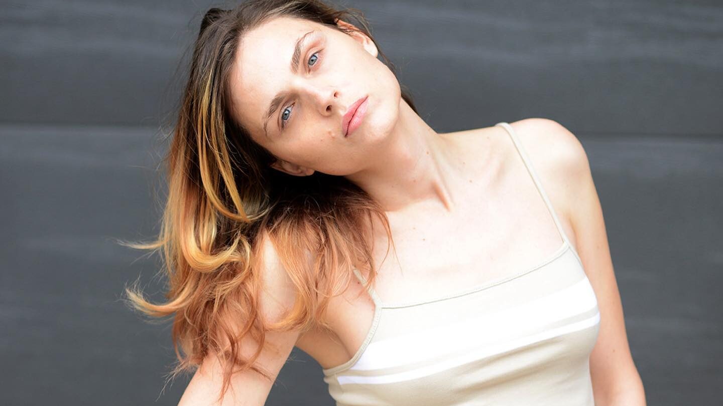 Andreja Pejić stands in front of a gray background, wearing a white spaghetti-strap crop top and high-waisted white jeans, her head tilted and hair falling over her shoulder.