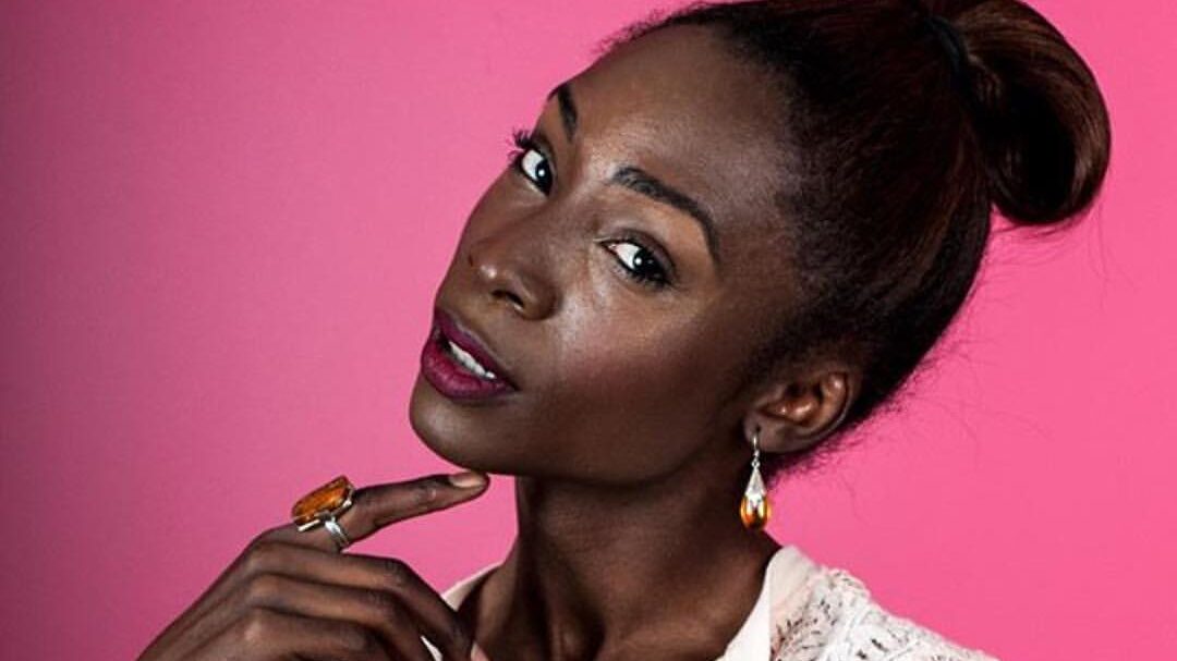 Angelica Ross poses in front of a pink background, wearing a white lace top and earrings, hand under chin.