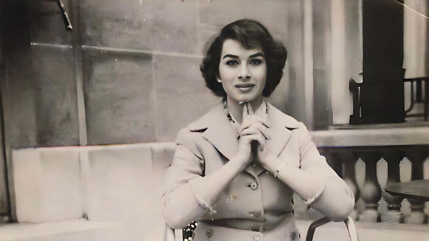 Black-and-white photo of April Ashley sitting on a chair, wearing a tailored suit and heels.