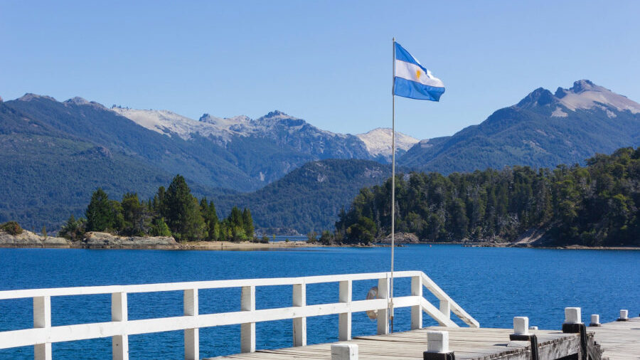 Drapeau argentin sur un ponton face à un lac bleu et des montagnes boisées en Patagonie sous ciel dégagé.