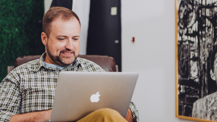 Bearded man in plaid shirt smiling while working on a silver laptop in a casual room with framed art on the wall.