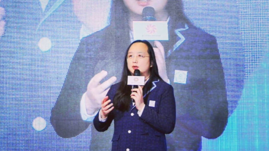 Audrey Tang speaks onstage holding a microphone with her image projected large behind.