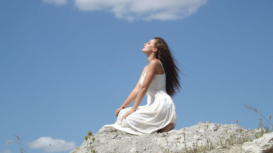 Mujer con un vestido blanco fluido, sentada en un terreno rocoso, respirando aire fresco.