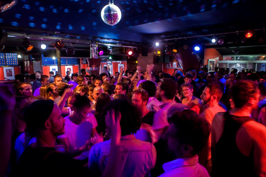 A crowded dance floor at La Boîte à Frissons nightclub in Paris, with LGBT+ people dancing under colorful disco lights.