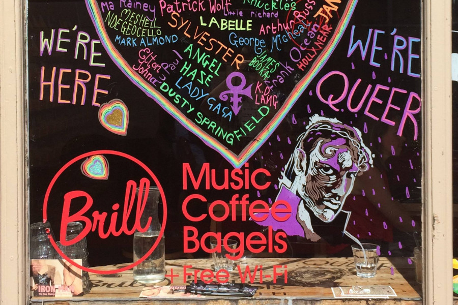 Café window decorated for Pride with rainbow heart listing queer music icons and “We’re here / We’re queer” slogans.