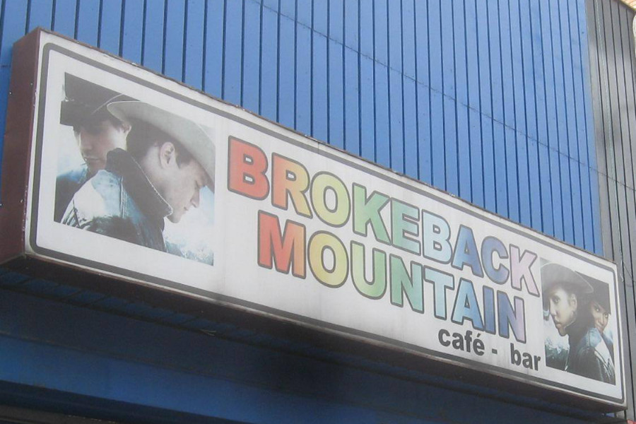 Letrero de Brokeback Mountain Café Bar que incluye texto con los colores del arcoíris e imágenes de la película en una pared azul exterior.