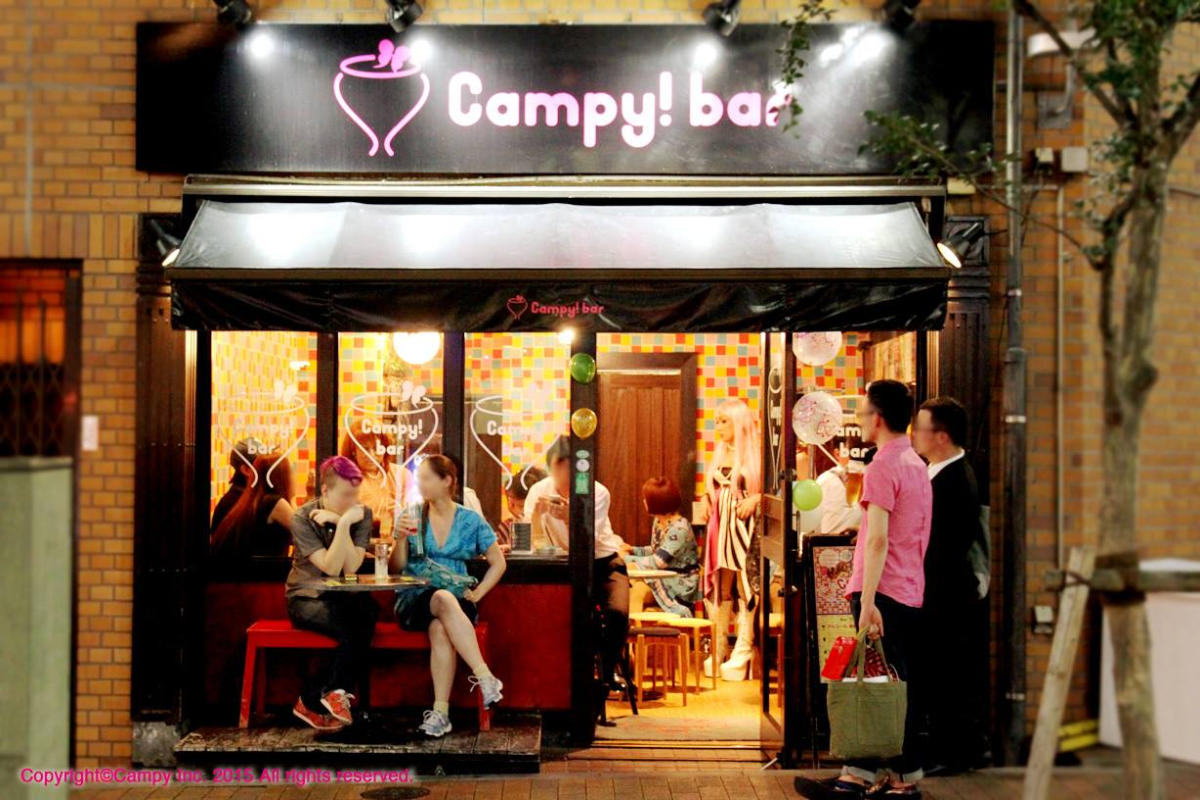 Street view of Campy! bar at night, showing a small crowd of people sitting and standing outside the brightly lit entrance.