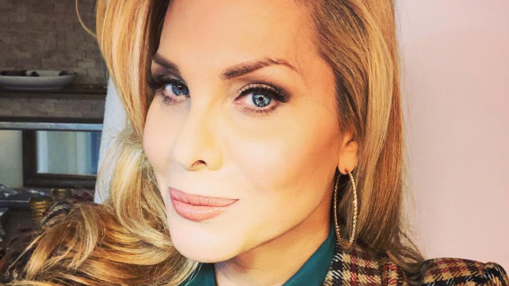 Candis Cayne smiling in a close-up selfie, wearing hoop earrings and a plaid jacket over a green blouse.