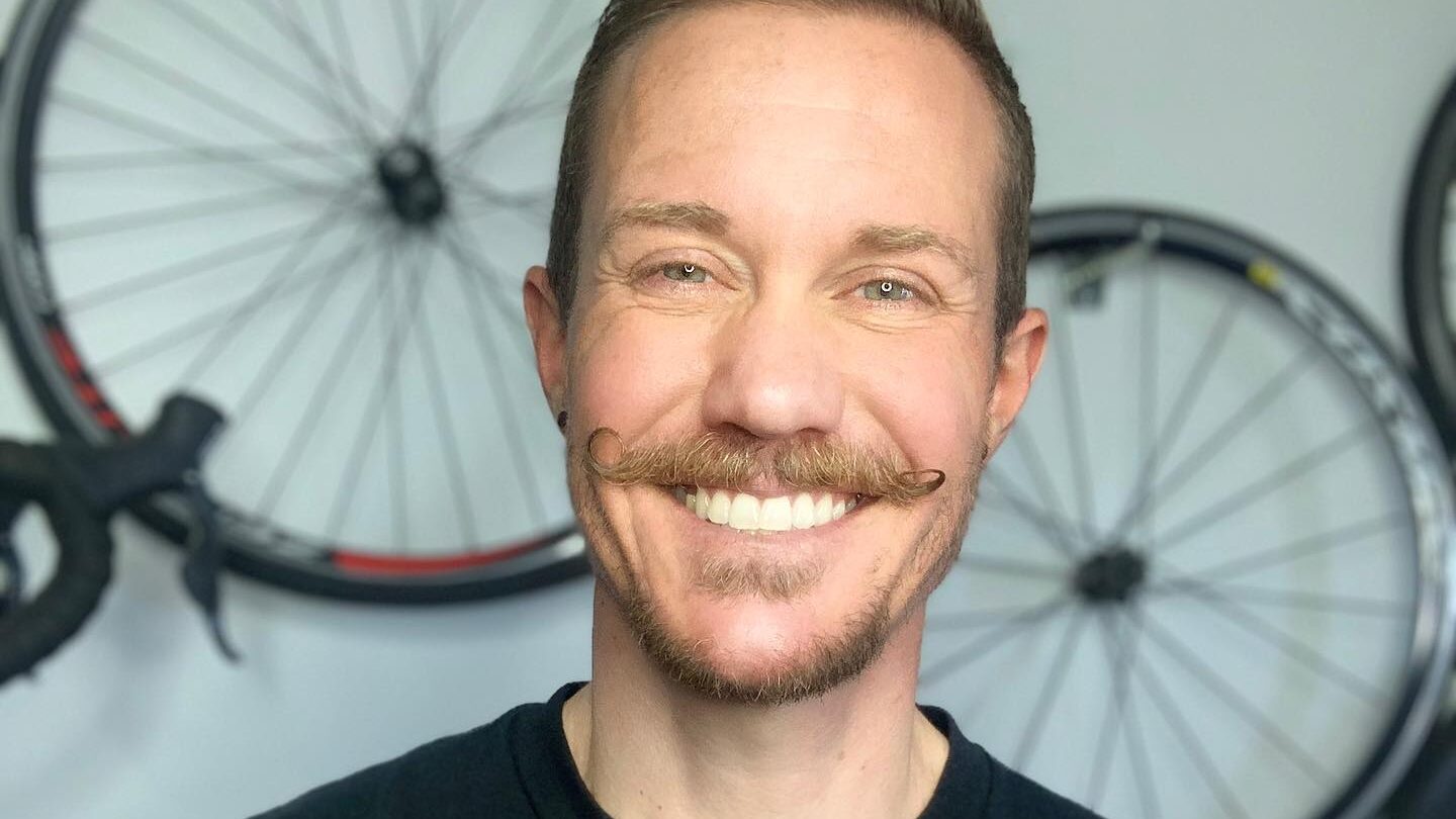 Chris Mosier sonríe frente a unas bicicletas, con una camiseta negra que dice “Activist Athlete” en casa.