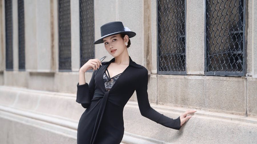 Fashionably dressed woman leaning on a stone wall, wearing a black outfit and holding sunglasses.