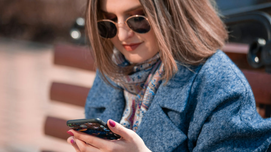 A woman in a blue coat and scarf sits on a wooden bench, wearing sunglasses while reading her phone screen.
