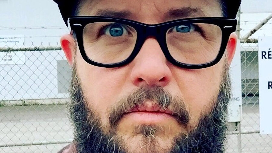 Cooper Lee Bombardier with beard, glasses, and cap, wearing a checkered shirt outdoors.