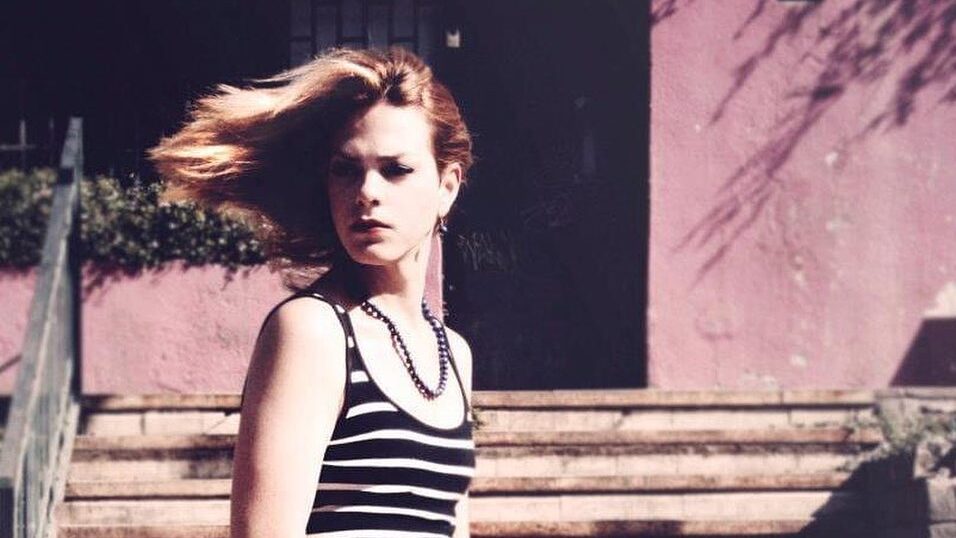 Young Daniela Vega seated on tiled stairs in striped dress, wind blowing her hair.