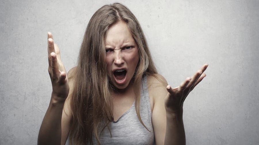 Woman with long hair yelling with hands raised in frustration, wearing a gray tank top against a plain background.