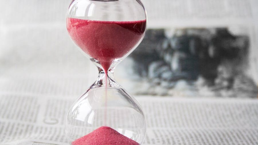 Glass hourglass with red sand flowing from top to bottom, placed on an open newspaper.
