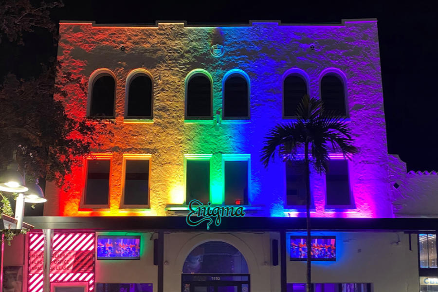 The exterior of Enigma St. Pete lit in rainbow colors, with arched windows and a palm tree at night.