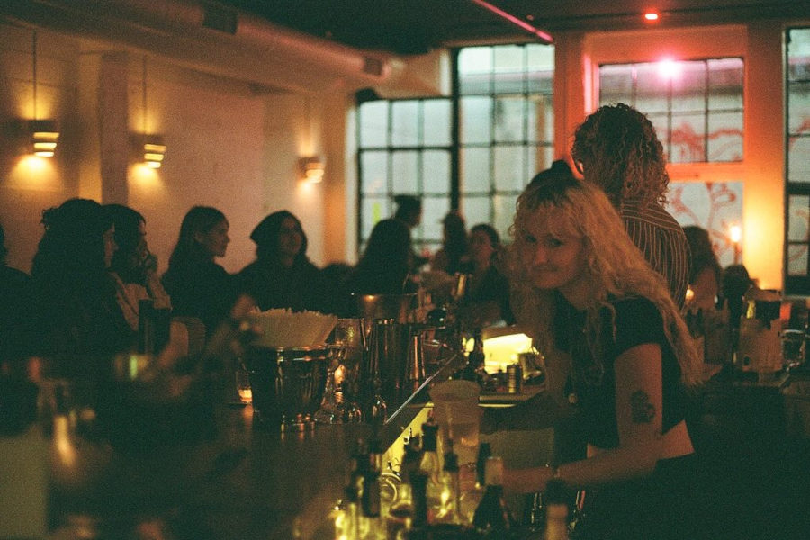 Inside Friends and Family Bar in New York City. A warmly lit bar scene shows guests chatting and relaxing. A smiling bartender stands behind the counter in a cozy, inclusive atmosphere.
