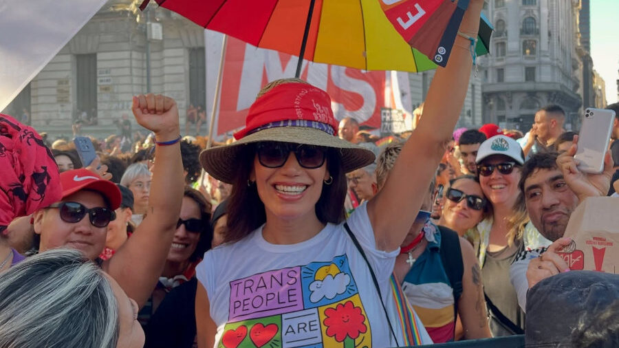 Florencia de la V souriante à Plaza de Mayo, entourée de manifestants et drapeaux arc-en-ciel.