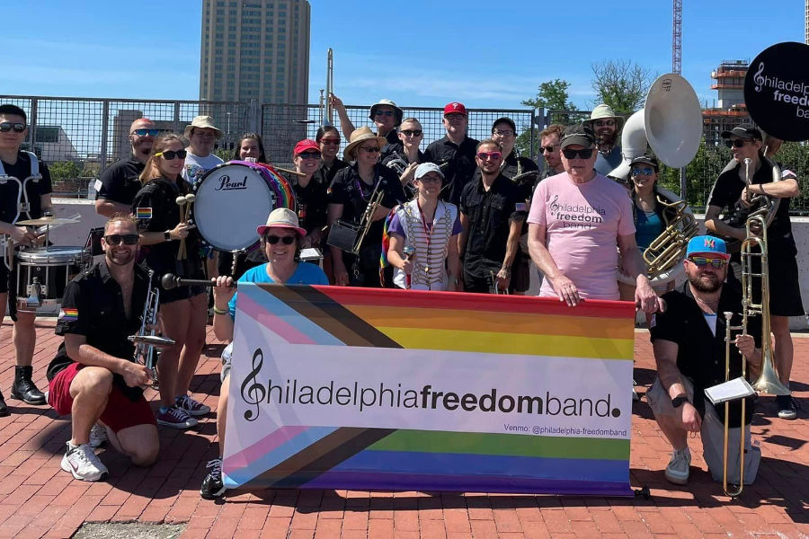 Members of the Philadelphia Freedom Band posing with instruments and a Pride banner on a brick path, city skyline behind.