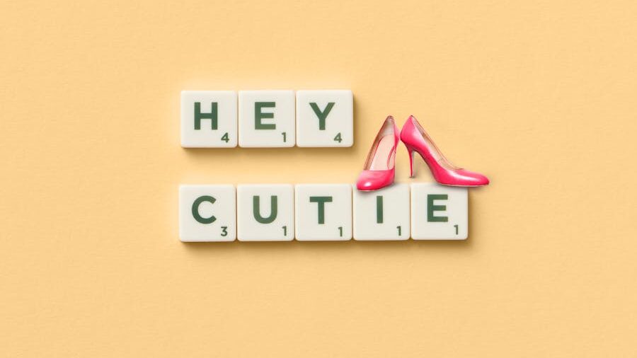 Scrabble tiles spell “HEY CUTIE” on a beige background, decorated with two pink high heel toy shoes.