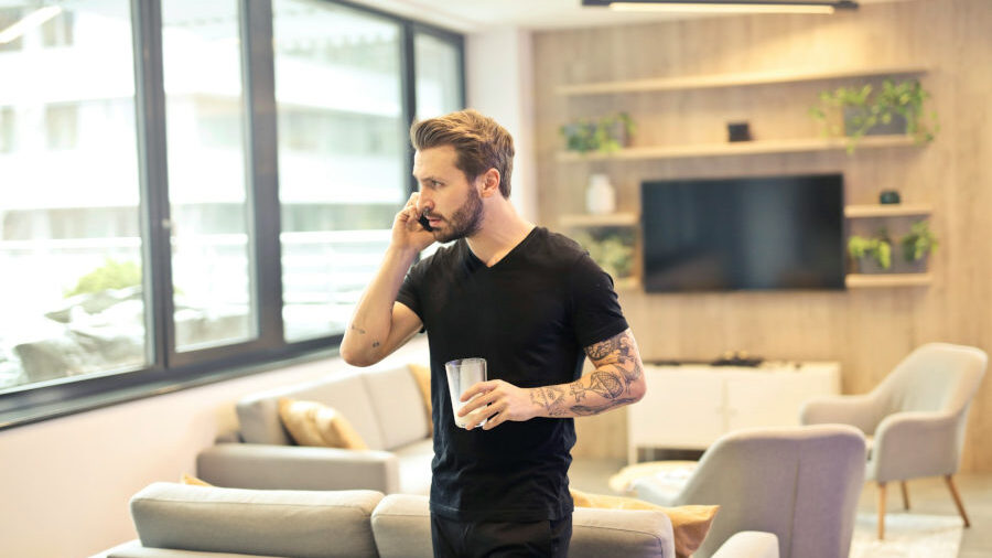 Tattooed man in a black shirt talks on the phone while holding a glass of water in a modern, sunlit apartment.