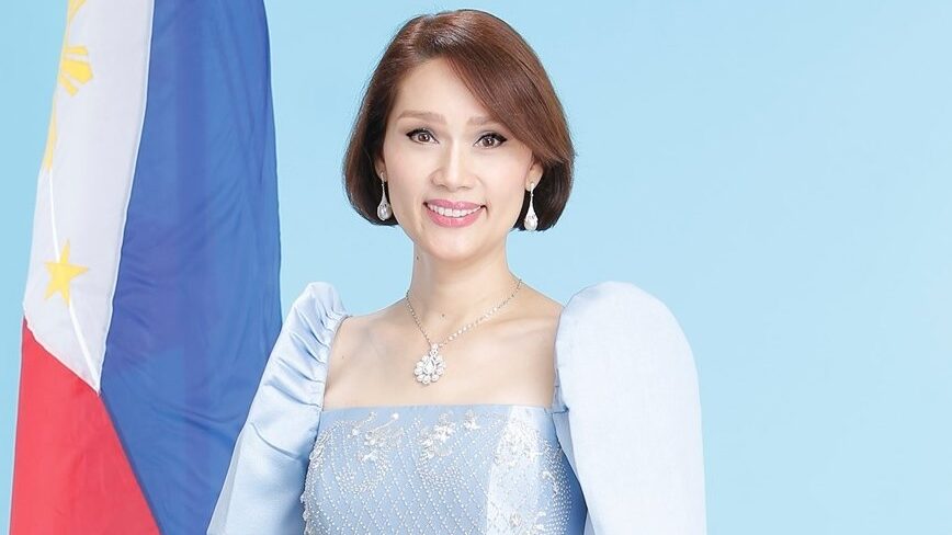 Geraldine Roman smiling in a light blue terno, standing in front of the Philippine flag.
