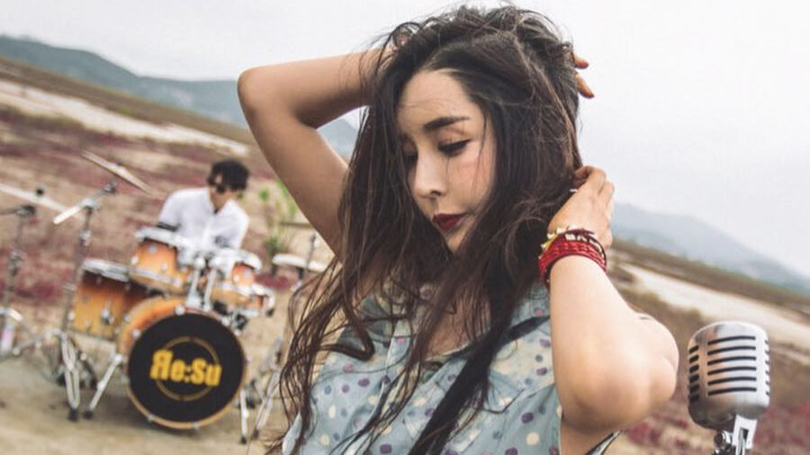 Harisu poses with a vintage microphone in front of a drummer, standing in a vast outdoor landscape with red grass.