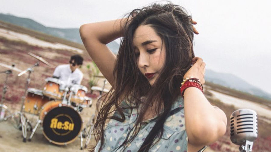 Harisu poses with a vintage microphone in front of a drummer, standing in a vast outdoor landscape with red grass.