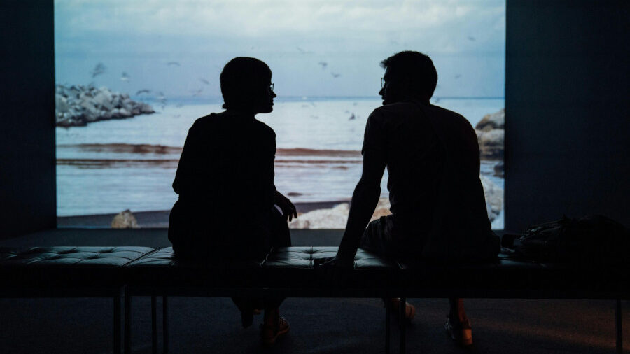 Two silhouetted figures sitting on a bench in front of a large screen displaying a coastal scene with ocean and rocks.
