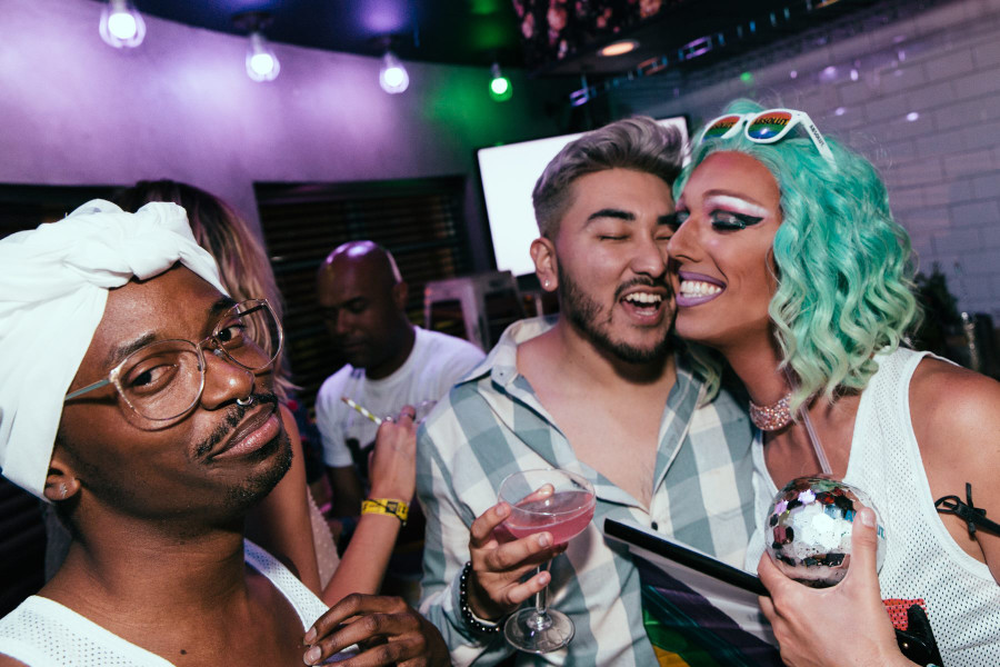 Drag performer with teal wig hugging a friend at a vibrant Pride celebration indoors.