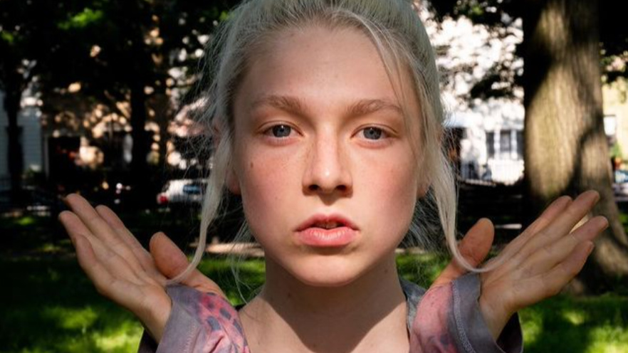 Hunter Schafer posando al aire libre con luz natural suave, usando guantes largos sin dedos y mirando con calma a la cámara.