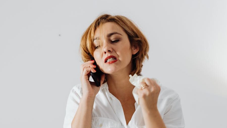 A woman in a white shirt talks on the phone with a distressed expression and clenched fist, holding a tissue.