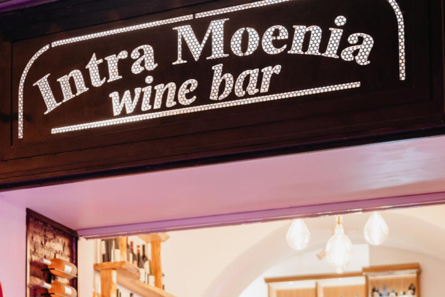 L’interno dell’Intra Moenia con l’insegna di legno, gli scaffali pieni di bottiglie e un barista dietro al bancone decorato con piastrelle azzurre.