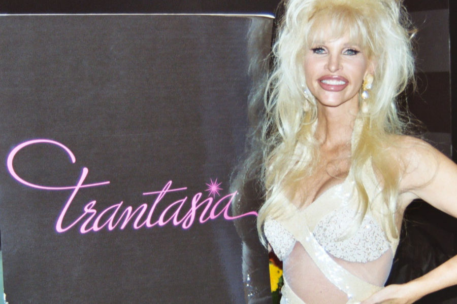 Jahna Steele smiling in a white dress beside a pink Trantasia sign, image from tribute article.