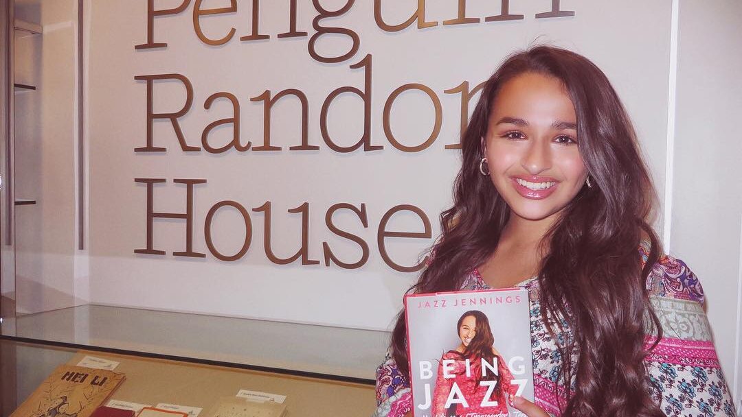 Jazz Jennings smiling while holding her memoir Being Jazz: My Life as a (Transgender) Teen in front of a Penguin Random House sign.