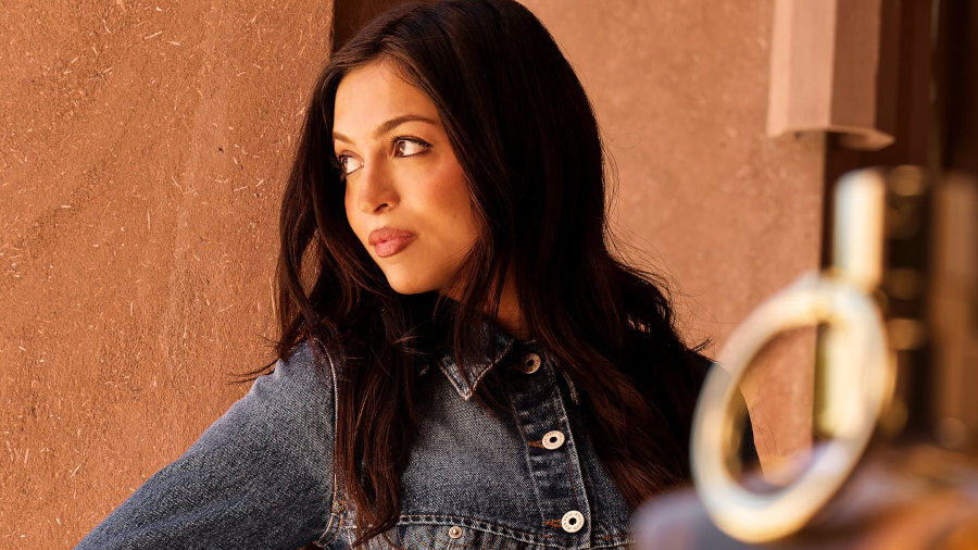 Josie Totah poses in a cropped denim jacket and jeans by Jean Paul Gaultier, leaning against a textured beige wall.