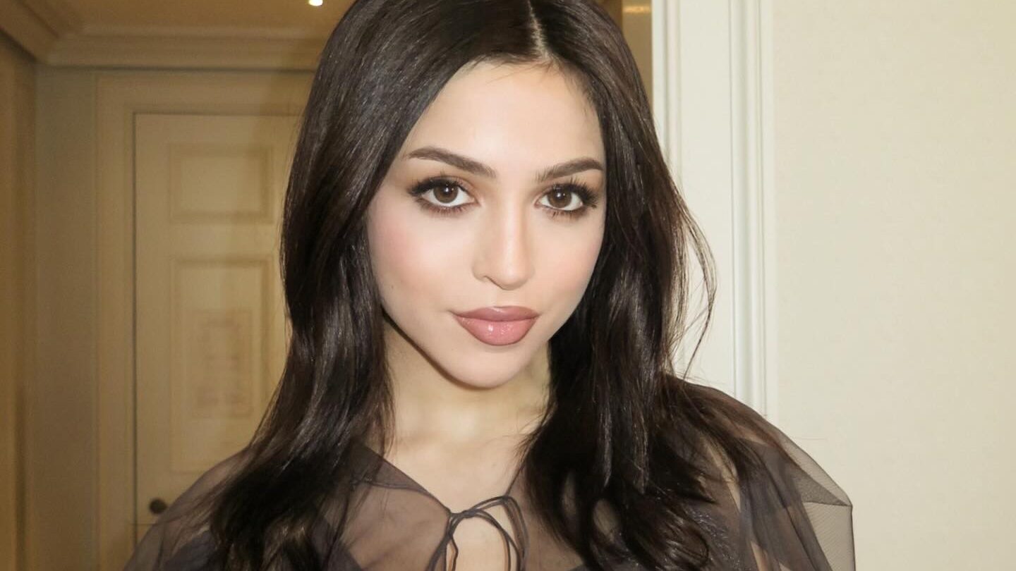 Josie Totah dans un couloir d’hôtel à Londres, en top noir transparent, maquillage et coiffure glamour.