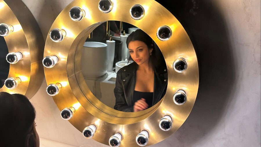 Josie Totah sits at a round lighted vanity mirror, reflection centered, with drink and phone on marble counter.