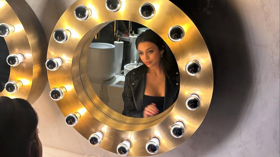 Josie Totah assise face à un miroir rond éclairé, reflet centré, verre et téléphone sur le comptoir en marbre.