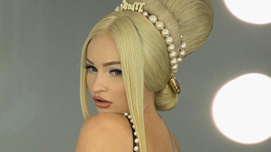 Kim Petras in a black pearl-trimmed dress with a dramatic retro beehive hairstyle, posing in a dressing room with lit vanity mirrors.