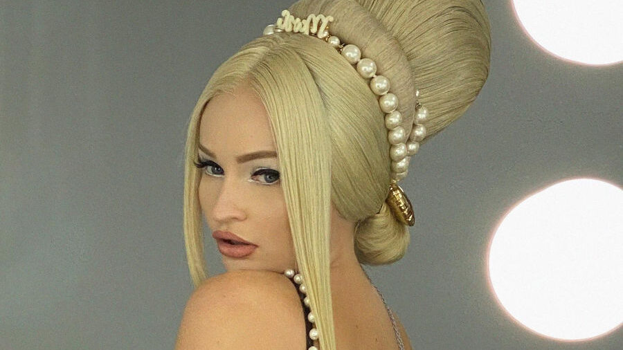 Kim Petras en robe noire bordée de perles, coiffée d’un chignon rétro spectaculaire, posant dans une loge devant un miroir éclairé.