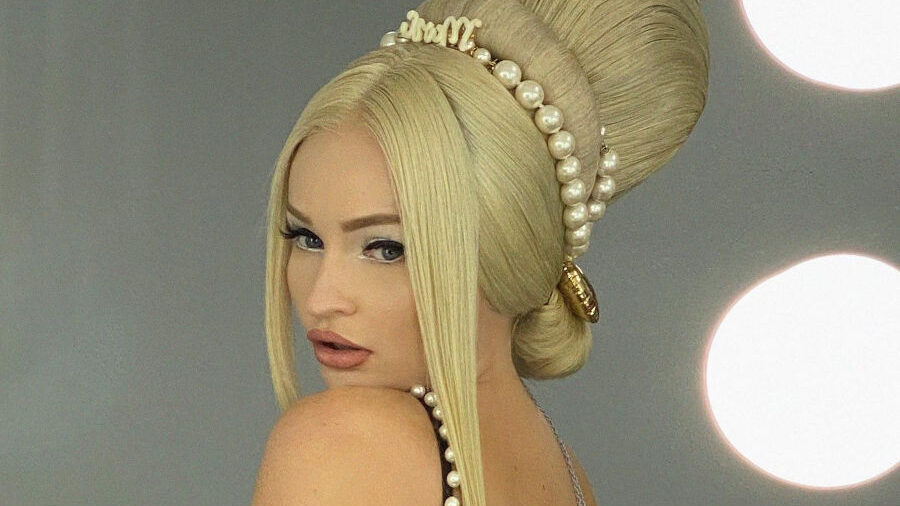 Kim Petras in a black pearl-trimmed dress with a dramatic retro beehive hairstyle, posing in a dressing room with lit vanity mirrors.