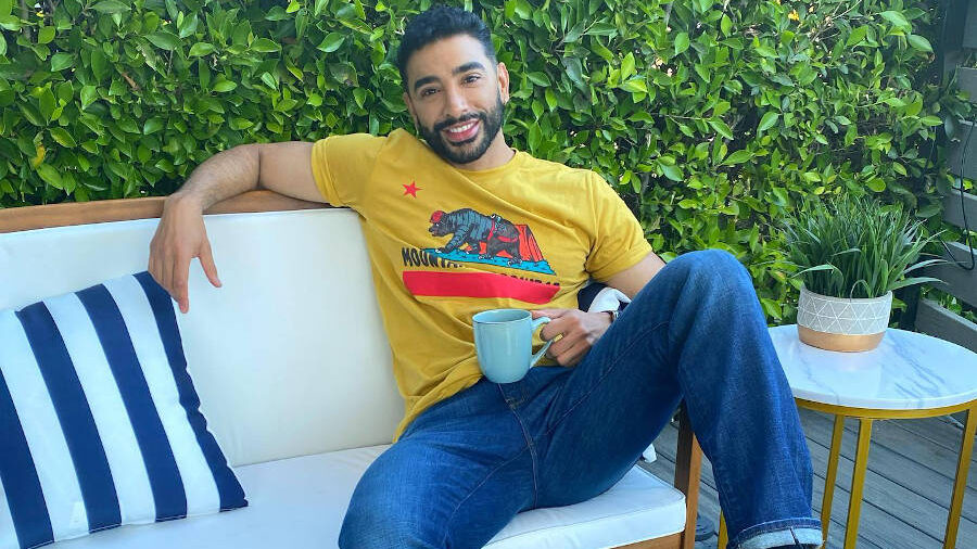 Laith Ashley sentado en un sofá blanco al aire libre con una taza de café, sonriendo de forma relajada.