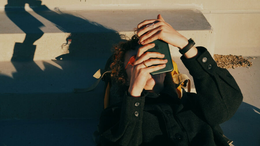 Person lying on sunlit steps, holding a smartphone above their face, wearing a dark coat and smartwatch.