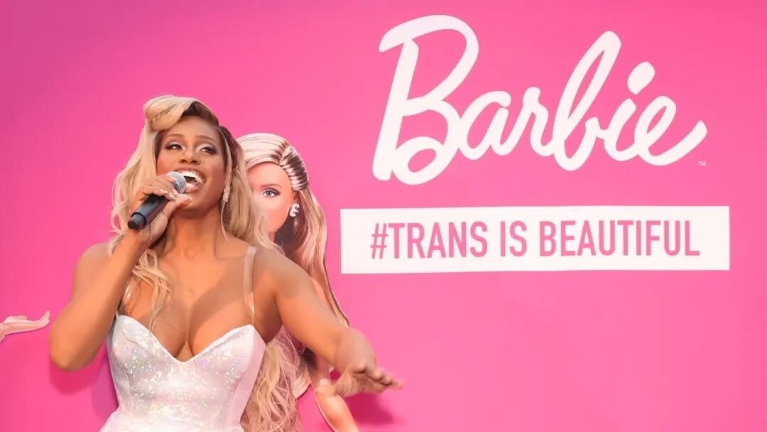 Laverne Cox poses joyfully in a sparkly white jumpsuit, microphone in hand, in front of a hot pink Barbie backdrop with her Barbie doll beside her. The bold text “#TRANS IS BEAUTIFUL” is displayed behind her, celebrating her groundbreaking collaboration with the iconic doll brand.