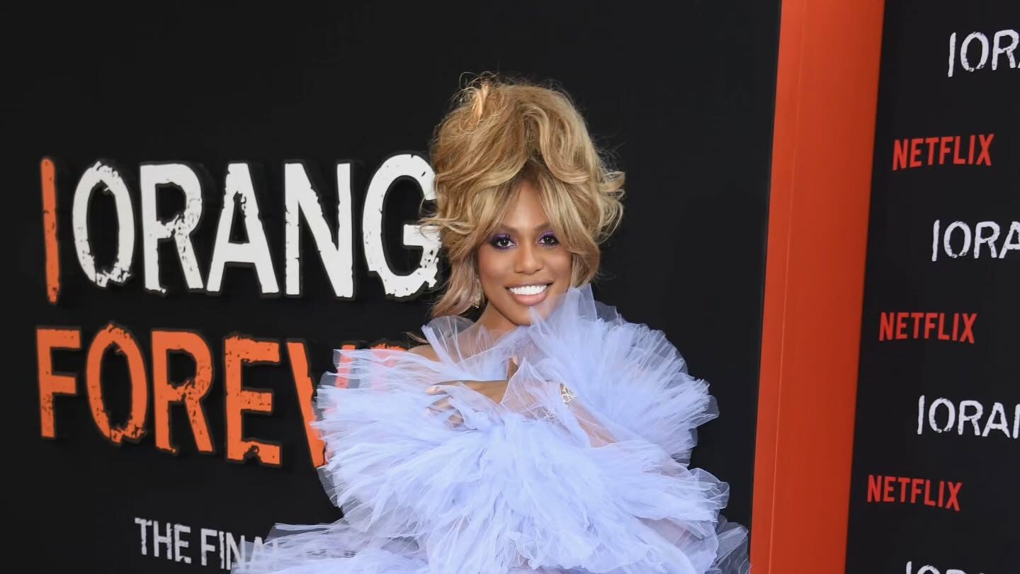 Laverne Cox pose dans une robe volumineuse lavande sur le tapis rouge de la célébration des 10 ans de la série Orange Is the New Black. Elle sourit devant un décor noir et orange de Netflix où l’on peut lire « ORANGE FOREVER ».