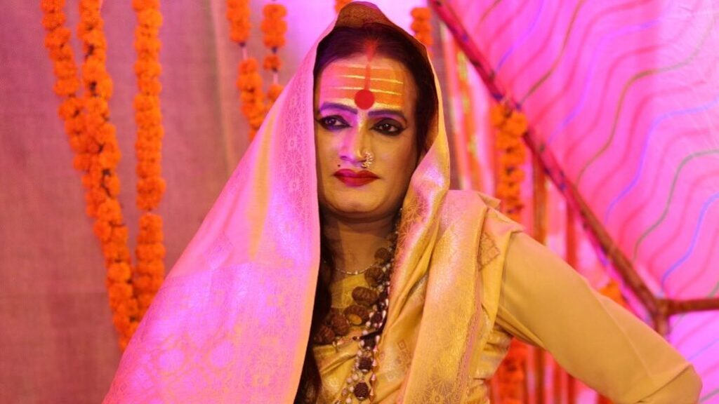 Laxmi Narayan Tripathi sentada con las piernas cruzadas, con un sari dorado y amarillo, bajo luces rosadas.