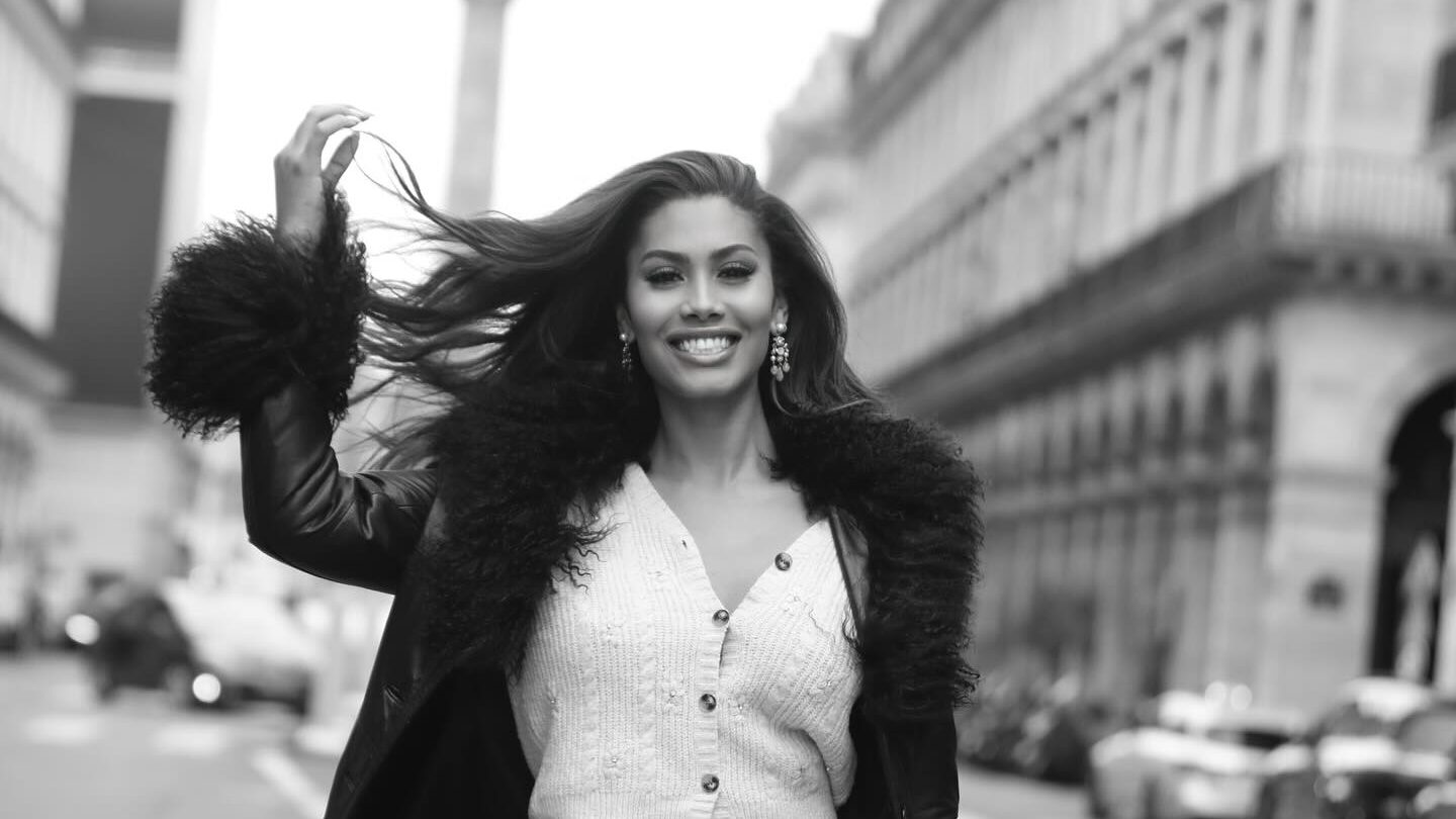 Leyna Bloom walking confidently in the middle of a street in Paris, wearing a fur-trimmed coat over a knit skirt set, holding a handbag, with Haussmannian architecture in the background.
